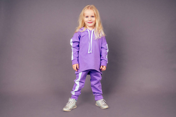 blonde blue-eyed little model girl in a purple sports suit posing in the studio on a black background.active childhood full length portrait violet sporty costume - Valokuva, kuva
