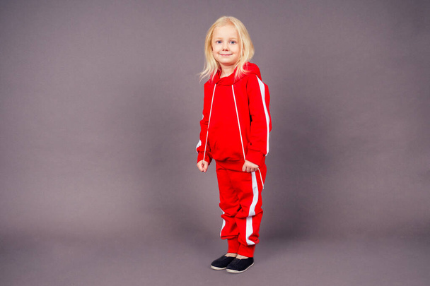 blonde blue-eyed little model girl in a red sports suit posing in the studio on a black background.active childhood full length portrait sporty costume hip-hop dancer - Фото, зображення
