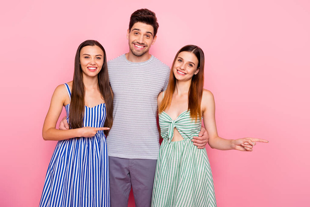 Portrait of cheerful man and two woman with long ginger foxy hair pointing at copy space wearing striped dress skirt t-shirt isolated over pink background - Zdjęcie, obraz