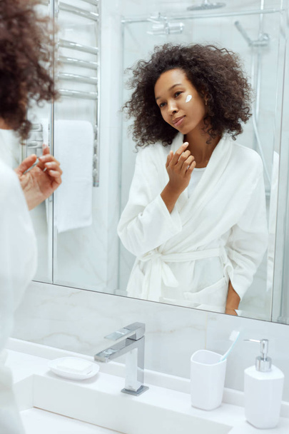 Skin care. Woman applying face cream looking in bathroom mirror - 写真・画像