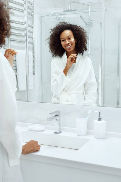 Skin care. Woman applying face cream looking in bathroom mirror - Fotó, kép