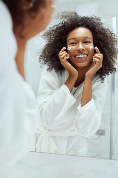 Skin care. Woman applying face cream looking in bathroom mirror - Фото, изображение