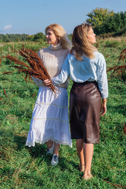 Portrait of two beautiful women in a field in late summer. Beautiful girls standing next to each other and holding bouquet made of dry brown plants. - Фото, зображення