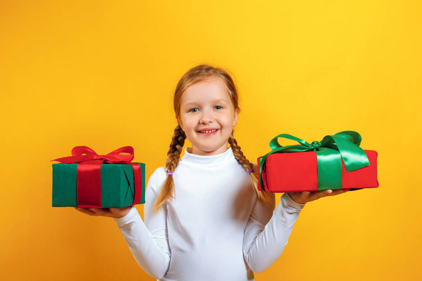 Portrait of a happy little girl in a white turtleneck on a yellow background. The child holds boxes with gifts in his hands - Foto, afbeelding