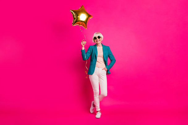 Вид в полный рост на красиво выглядящую привлекательную жизнерадостную светловолосую даму с воздушным шариком в руках, наслаждающуюся свободным временем на ярком светящемся ярко-розовом цветовом фоне фуксии
 - Фото, изображение