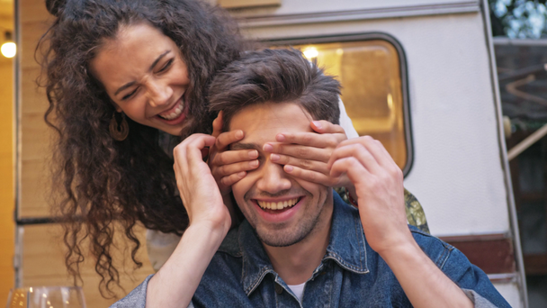 Attractive cheerful young brunette woman with curly hair making surprise to her boyfriend and closing his eyes with hands while spending time near the trailer - Footage, Video