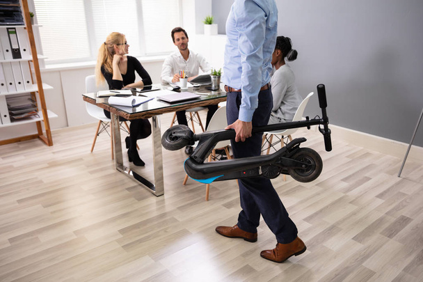 Businessperson Carrying Folded Electric Scooter In Front Of His Colleagues At Office - Foto, imagen