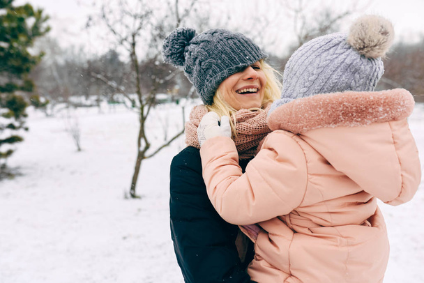 Happy mother holding her child, smiling broadly and havinf fun during walking in the park on snowing day. Young woman and her daughter playing together. Christmas mood. Childhood - Photo, image