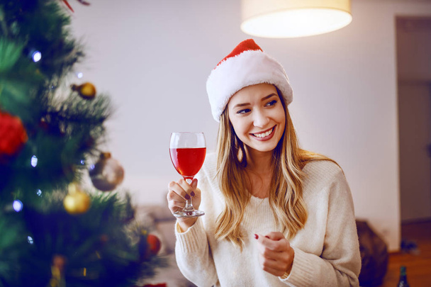 Vidám, kaukázusi szőke nő mikulás sapkával a fején mosolyog és borral koccint, miközben a karácsonyfa mellett áll a nappaliban.. - Fotó, kép