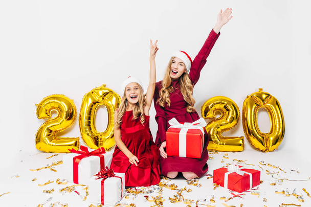a happy mother and little daughter in Santa hats, having fun sitting on a white background with Christmas gifts in their hands - Fotó, kép