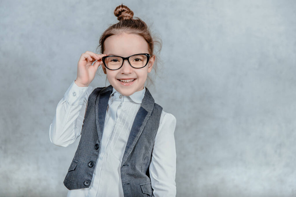 Business little cute baby girl in glasses on gray background. During this, she holds his glasses and looks at the camera. - 写真・画像