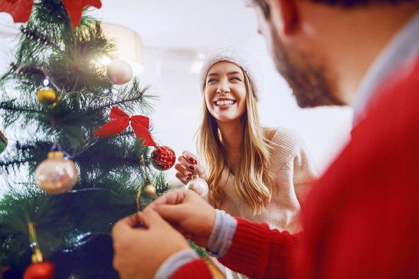 Rozkošný kavkazský pár zdobí vánoční stromeček. Oba mají na hlavách Santa Klobouky. Interiér obývacího pokoje. - Fotografie, Obrázek