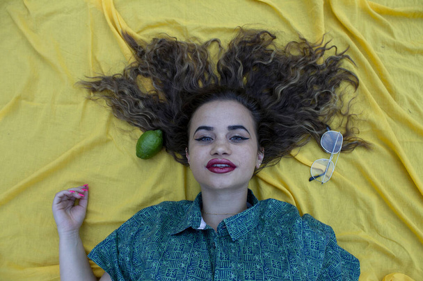 Mladá žena ležící na žluté látce jako pozadí. S kudrnatými hnědými vlasy. - Fotografie, Obrázek