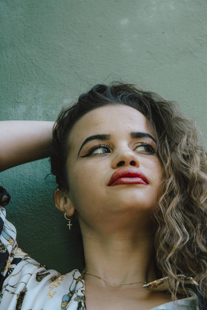 Portrait of young woman with urban clothes, with curly hair. On a green wall in the background, made up to party. - Foto, afbeelding