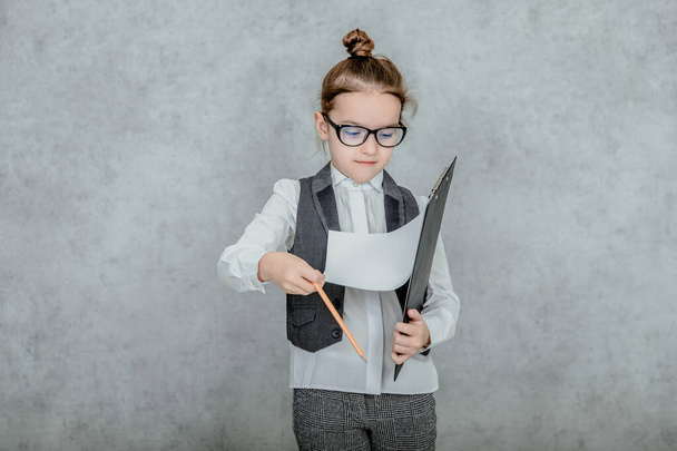 Little girl with a folder in her hands on a gray background. Dressed up as a business lady and glasses. Viewing business documents. - Photo, Image