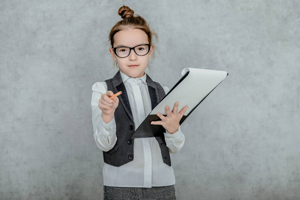 Little girl with a folder in her hands on a gray background. Dressed up as a business lady and glasses. Looking at the camera, bewildered, showing a pencil to the camera. - Foto, Bild