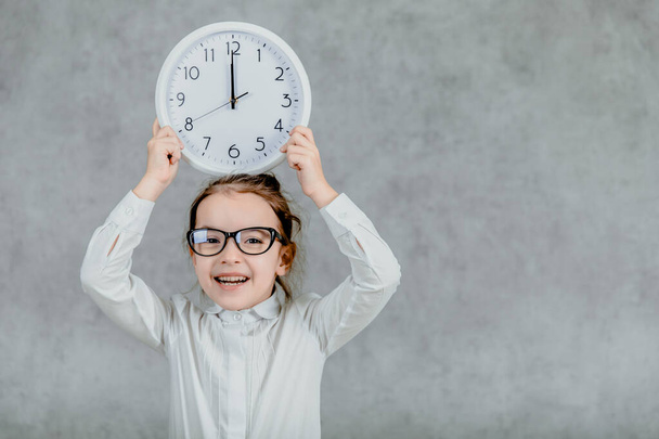 Beautiful little girl with a clock on a gray background. Lift his hands up with the clock. Emotional - Φωτογραφία, εικόνα
