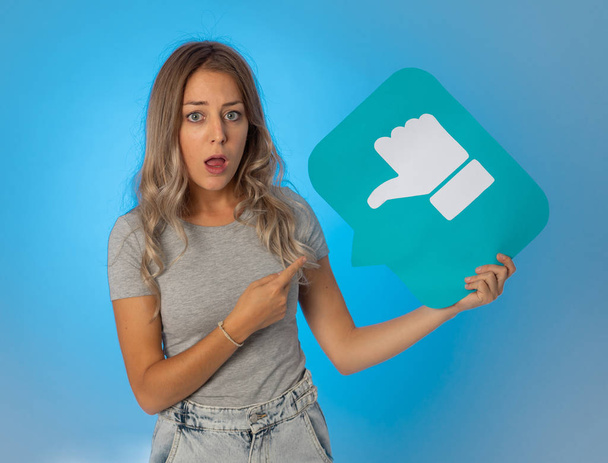Attractive unhappy young woman holding social media symbol of dislike feeling sad and rejected in internet obsession, social network notification icons, cyber bullying and technology communication. - Фото, зображення