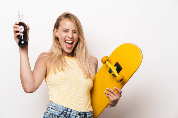 Cheery amazing young pretty woman posing isolated over white wall background holding skateboard drinking soda. - Fotografie, Obrázek