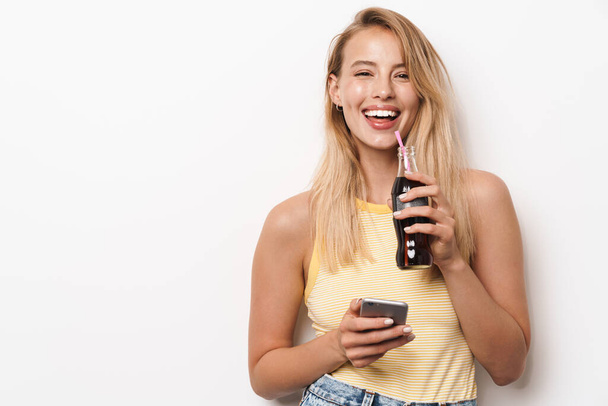 Amazing young pretty woman posing isolated over white wall background using mobile phone drinking soda. - Zdjęcie, obraz
