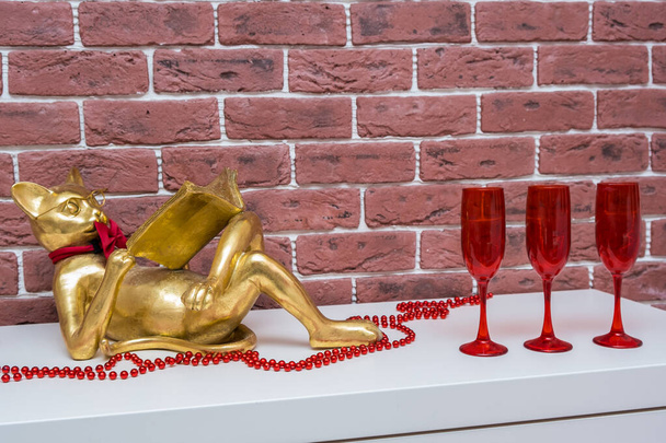 Золотий кіт читає книгу лежить на білій поверхні з червоним бісером і червоними келихами для вина, інтер'єр вітальні
 - Фото, зображення