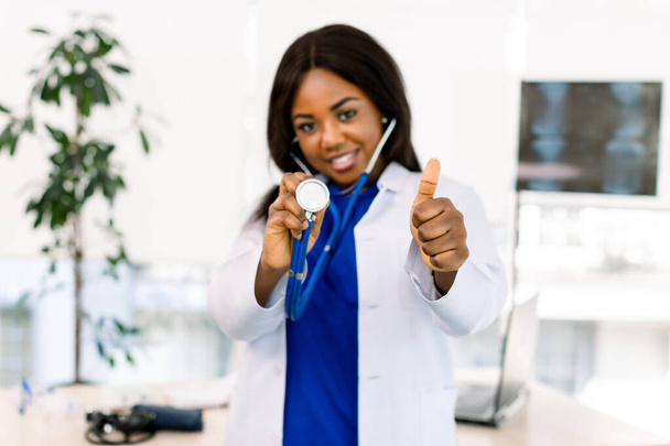 Closeup portrait of friendly, smiling confident African woman healthcare professional in lab coat, stethoscope, gesturing thumb up. Isolated hospital clinic background. - 写真・画像