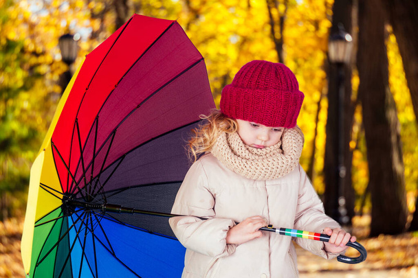 秋の公園のグレーのジャケットに虹色の傘を持つ悲しい面白い美しいブロンドの子供。屋外で遊んでいる女の子の子供。9月の森の中を歩く黄色い木の葉 - 写真・画像