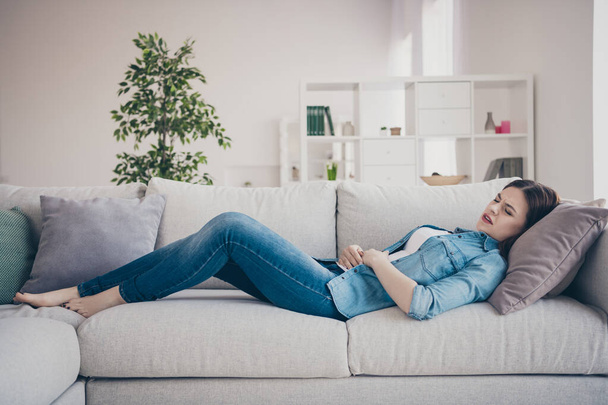 Profile photo of pretty lady suffering from pms holding hands on hurted belly lying sofa trying to relax wearing jeans outfit apartment indoors - Foto, Imagem