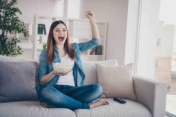 Photo of amazing lady watching long waited sport game eating popcorn raising arm up supporting team sitting comfy sofa wearing jeans clothes apartment indoors - Foto, immagini