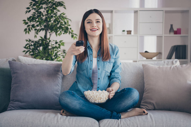 Photo of amazing lady going to watch favorite humor tv show holding popcorn plate on crossed legs sitting comfy sofa wearing jeans clothes apartment indoors - Фото, изображение