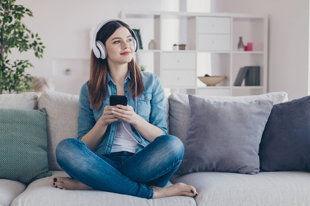 Photo of pretty dreaming lady with telephone in hands turning on cool modern song in earflaps sitting comfy sofa wearing jeans clothes apartment indoors - Foto, immagini