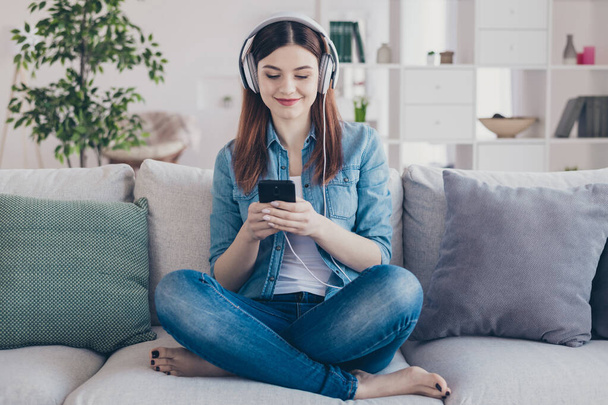 Photo of amazing lady with telephone in hands turning on cool modern song in earflaps sitting comfy sofa wearing jeans clothes apartment indoors - Photo, image