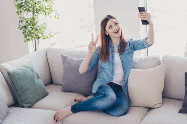 Photo of pretty lady with telephone in hands making selfies showing v-sign symbol sitting comfy on sofa wearing jeans clothes apartment indoors - Foto, imagen