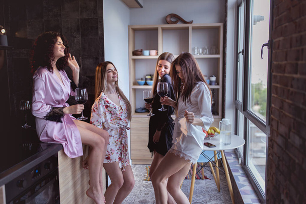 Buen humor hermosas damas disfrutando del tiempo juntos mientras celebran la despedida de soltera en casa en un estudio moderno sosteniendo copas de vino y moviéndose divertido delante de la cámara
 - Foto, Imagen