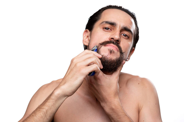 Pohledný muž s knírkem používá elektrický holicí strojek k zastřihování vousů, které se nacházejí na bílém pozadí. Koncepce ranní léčby a holení. Čas zastřihnout vousy. Ranní rutina - Fotografie, Obrázek
