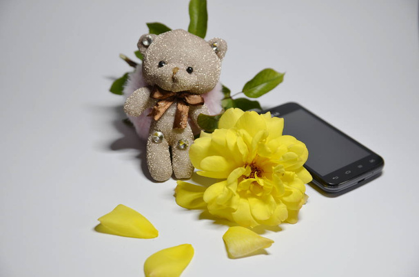 Egy fehér háttérben egy vicces kis maci, egy sárga friss rózsa és egy telefon.. - Fotó, kép
