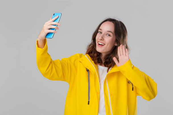 Mooie brunette vrouw in een gele regenjas selfie maken met smartphone geïsoleerd over grijze achtergrond. Gaan wandelen op een regenachtige koude dag - Foto, afbeelding