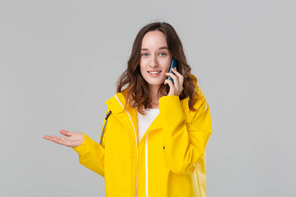 Pretty brunette woman in a yellow raincoat looking happy talking on the mobile phone isolated over grey background. Concept of communication. - Photo, Image