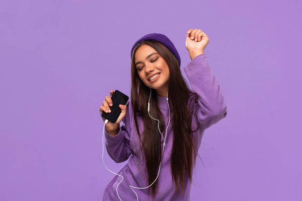 Pretty african-american girl in a hat and sweater listening to music on a mobile phone with wired headphones and dancing isolated over purple background. Enjoying life. - Photo, Image