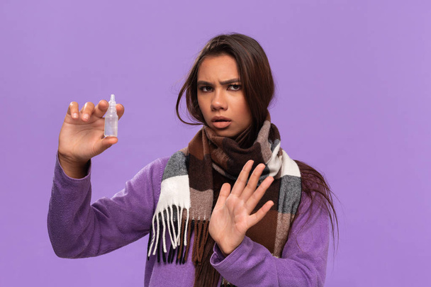 Досить афро-американська дівчина в светрі і затишний перевірений шарф з використанням носового спрею отримує лікування для бігу носа, що стоїть ізольовано на фіолетовому фоні. Застуда взимку
. - Фото, зображення