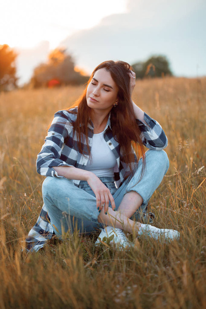 Beautiful young woman sitting on the grass at sunset. Eyes closed. - Φωτογραφία, εικόνα
