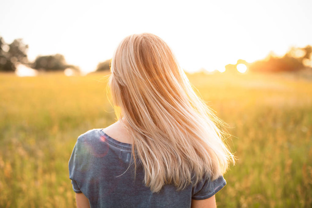 Young woman with blonde hair standing on the field and looking on the sunset - Valokuva, kuva