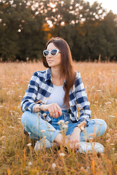 Young woman in plaid shirt and sunglasses is sitting on the dry grass - Fotoğraf, Görsel