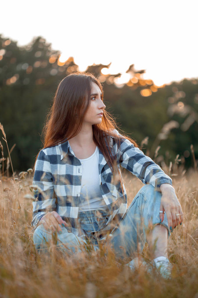 Beautiful young woman in plaid shirt sitting on the grass at sunset. Profile - Foto, Bild