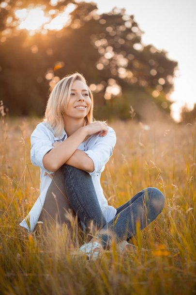 Young beautiful woman sitting on grass and smiles at sunset - Foto, Imagen