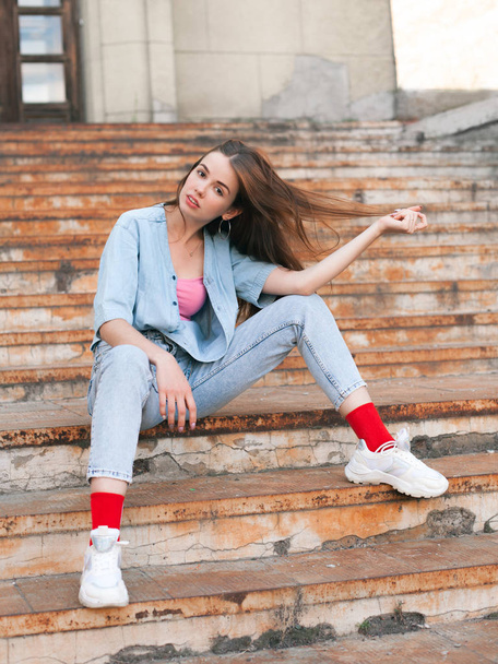 Beautiful young woman in jeans sitting on the old rusty stairs - Zdjęcie, obraz