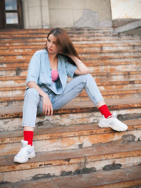 Beautiful young woman in jeans sitting on the old rusty stairs - Фото, изображение
