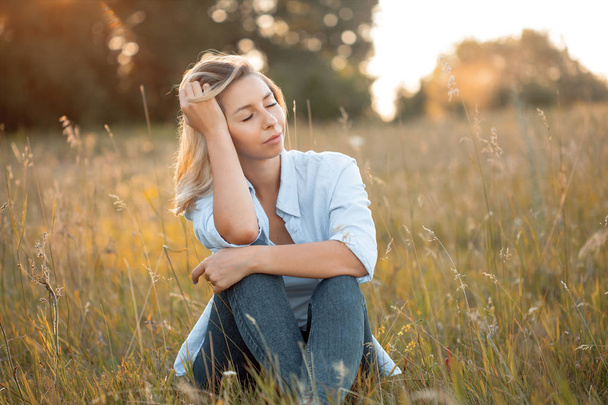 Young woman in blue shirt sitting on the green field under sunlight. Eyes closed - Photo, Image