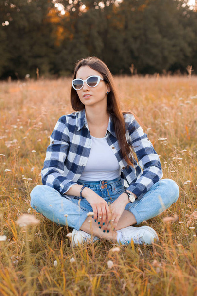 Beautiful young woman in plaid shirt and sunglasses is sitting on the dry grass - Foto, imagen