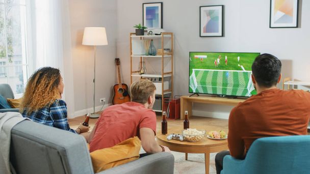 Trzech fanów sportu siedzi na kanapie w salonie i ogląda ważny mecz piłki nożnej w telewizji, wiwatując swojej drużynie, czekając na cel, który przynosi zwycięstwo ich drużynie. - Zdjęcie, obraz